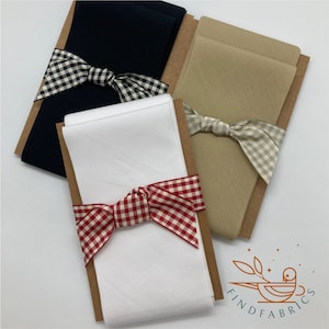 Könnte beinhalten: Drei Stoffmuster, die in braunes Papier eingewickelt und mit Vichy-Bändern gebunden sind. Die Muster sind weiß, beige und marineblau. Das weiße Muster ist mit einem rot-weißen Vichy-Band gebunden. Das beige Muster ist mit einem grau-weißen Vichy-Band gebunden. Das marineblaue Muster ist mit einem schwarz-weißen Vichy-Band gebunden. Die Verpackung ist mit "Find Fabrics" beschriftet.