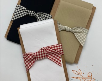 May include: Three fabric swatches wrapped in brown paper and tied with gingham ribbon. The swatches are white, beige, and navy blue. The white swatch is tied with a red and white gingham ribbon. The beige swatch is tied with a gray and white gingham ribbon. The navy blue swatch is tied with a black and white gingham ribbon. The packaging is labeled "Find Fabrics".
