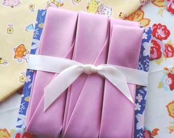 Puede incluir: Un paquete de cuatro cintas de algodón rosa, atadas con una cinta de satén blanco. La cinta está sobre un fondo de tela floral.