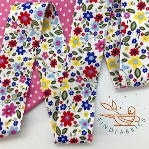 May include: White fabric with a floral print featuring red, blue, yellow, and pink flowers. The fabric is folded in half and the edges are visible. The fabric is on a pink polka dot background.