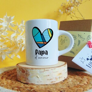 Peut inclure: Mug en céramique blanche avec un motif de cœur bleu, jaune et orange. La tasse dit "Papa d'amour" en texte noir.