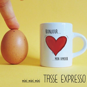Op de afbeelding: Witte espressokopje met een rood hart en de tekst "Bonjour... mon amour" in zwart. Het kopje staat op een gele ondergrond naast een bruin ei.