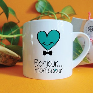 Op de afbeelding: Witte keramische mok met een turquoise hartontwerp. Het hart heeft een glimlachend gezicht en een zwarte strik. De tekst "Bonjour... mon coeur" is onder het hart gedrukt.