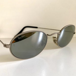 Puede incluir: Gafas de sol Ray-Ban plateadas con lentes ovalados y montura plateada. Las gafas tienen un acabado espejado.