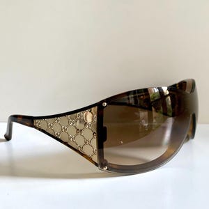 May include: Brown shield sunglasses featuring a wrap-around lens design with a gradient tint. The temples display a gold-toned metal panel embossed with a repeating interlocking G logo pattern, transitioning into a dark brown tortoiseshell frame.