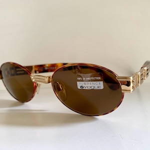 Może przedstawiać: Okulary przeciwsłoneczne w kształcie owalu, brązowe z złotymi elementami, z oprawką w kolorze imitującym pancerzyk żółwia. Okulary mają brązowe szkła i etykietę z napisem "100% UV Protection Florence Vogue".