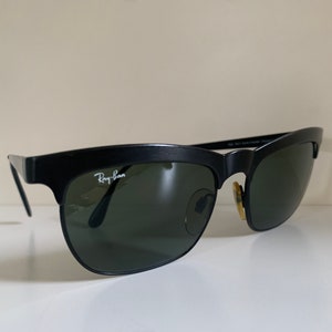 May include: Black Ray-Ban sunglasses with a classic design. The sunglasses have a matte black frame, dark green lenses, and the Ray-Ban logo in white on the left lens. The sunglasses are a timeless accessory.