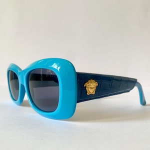 May include: Blue and black sunglasses with a gold Medusa head logo on the side. The sunglasses have a rounded, rectangular shape.