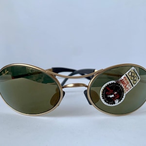 May include: Gold-framed sunglasses with oval lenses and a black bridge. The lenses are a dark green color, and a sticker with text and Olympic rings is affixed to one lens. The sunglasses are displayed against a white background.