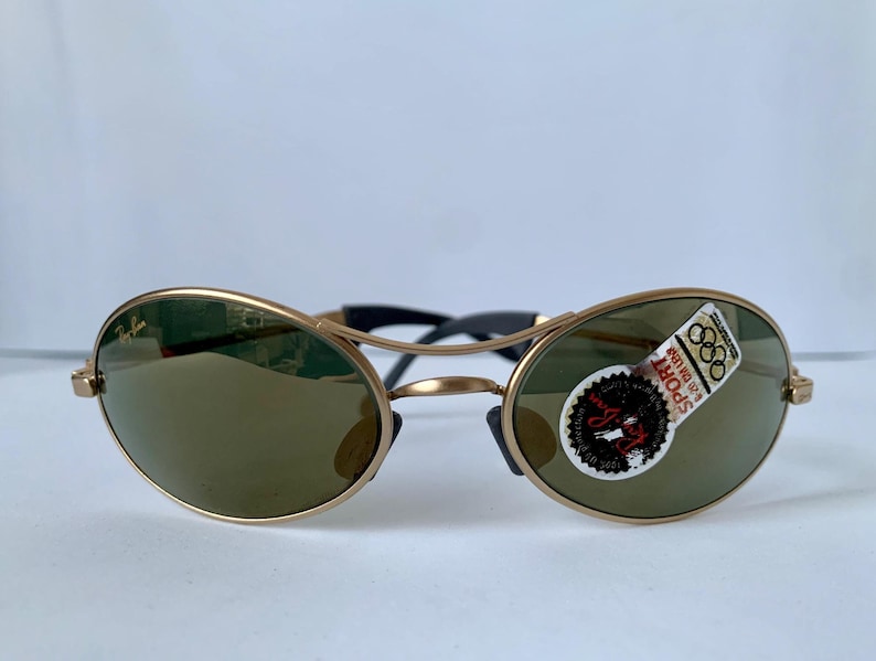 May include: Gold-framed sunglasses with oval lenses and a black bridge. The lenses are a dark green color, and a sticker with text and Olympic rings is affixed to one lens. The sunglasses are displayed against a white background.