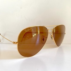 May include: Gold-framed aviator sunglasses with brown lenses. The sunglasses have a double bridge and the Ray-Ban logo is visible on the upper left lens. The temples are thin and gold-coloured.