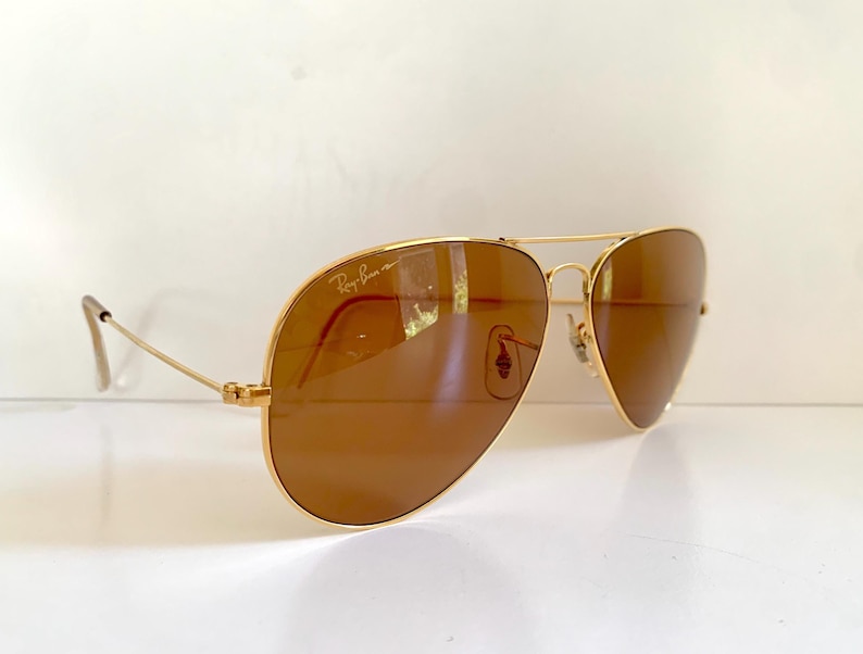 May include: Gold-framed aviator sunglasses with brown lenses. The sunglasses have a double bridge and the Ray-Ban logo is visible on the upper left lens. The temples are thin and gold-coloured.