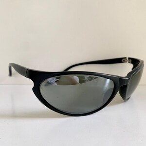 May include: Black Ray-Ban sunglasses featuring a wraparound frame design with dark, reflective lenses. The brand logo is printed in white on the upper corner of the right lens, and a small etched logo is visible on the lower edge of the same lens.