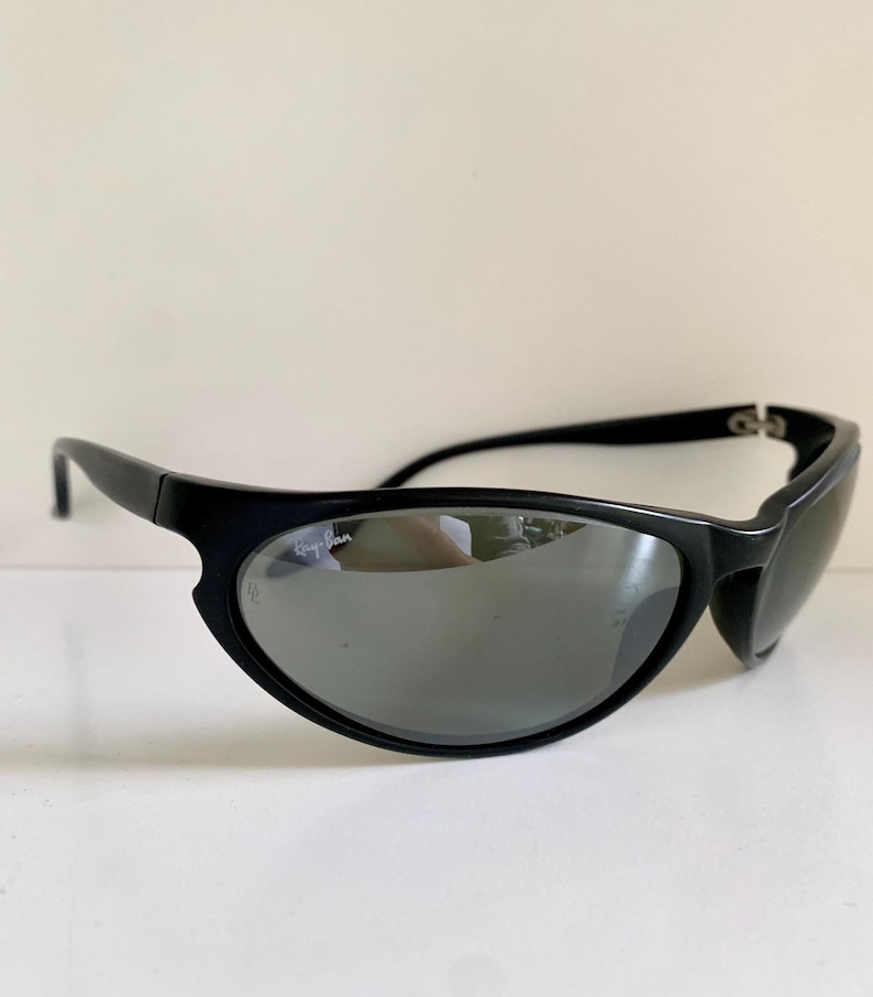 May include: Black Ray-Ban sunglasses featuring a wraparound frame design with dark, reflective lenses. The brand logo is printed in white on the upper corner of the right lens, and a small etched logo is visible on the lower edge of the same lens.