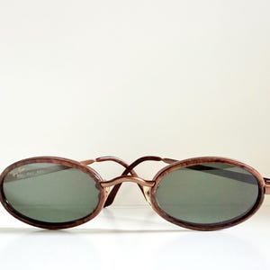May include: Oval sunglasses with a brown tortoiseshell frame and dark green lenses. The sunglasses have a thin, metal bridge and arms. The design is vintage-inspired and classic.