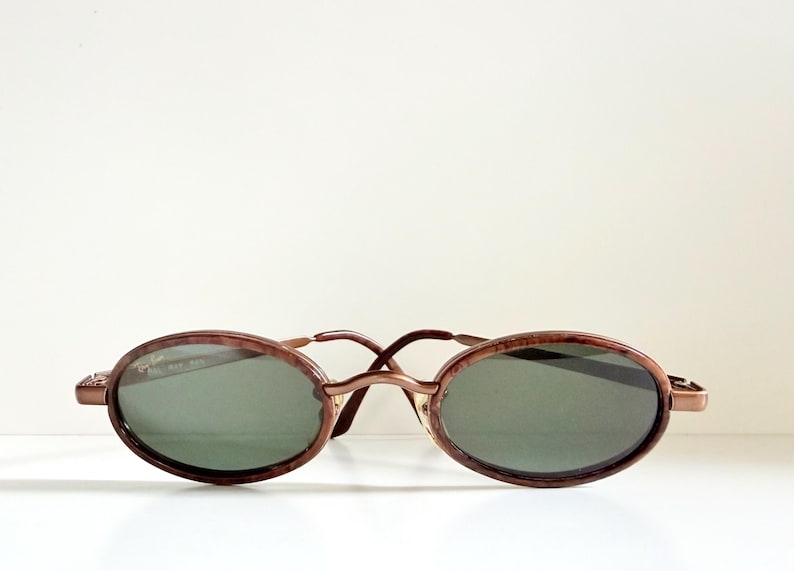 May include: Oval sunglasses with a brown tortoiseshell frame and dark green lenses. The sunglasses have a thin, metal bridge and arms. The design is vintage-inspired and classic.