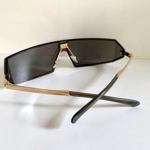 Peut inclure: Lunettes de soleil sans monture noires avec des branches dor&eacute;es. Les lunettes ont une forme g&eacute;om&eacute;trique et des verres fonc&eacute;s. Les branches sont dor&eacute;es pr&egrave;s de la monture et passent &agrave; une couleur plus fonc&eacute;e aux embouts. Sur une surface blanche.