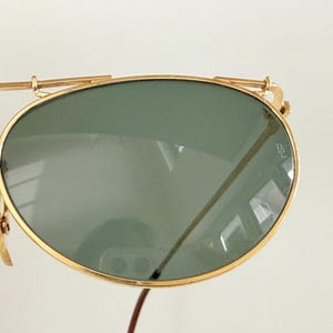 K&ouml;nnte beinhalten: Nahaufnahme einer goldgerahmten Sonnenbrille mit gr&uuml;nen Gl&auml;sern. Die Sonnenbrille hat ein klassisches Aviator-Design mit Doppelsteg und kleinem Logo auf dem Glas. Der Rahmen ist goldfarben gl&auml;nzend.