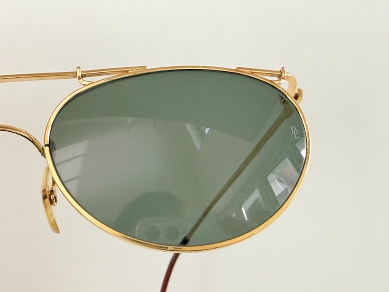 K&ouml;nnte beinhalten: Nahaufnahme einer goldgerahmten Sonnenbrille mit gr&uuml;nen Gl&auml;sern. Die Sonnenbrille hat ein klassisches Aviator-Design mit Doppelsteg und kleinem Logo auf dem Glas. Der Rahmen ist goldfarben gl&auml;nzend.