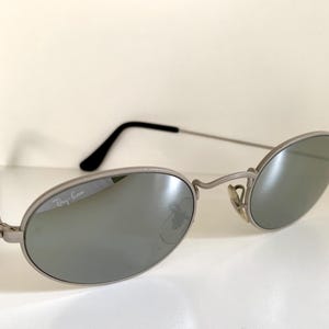 Puede incluir: Gafas de sol Ray-Ban plateadas con lentes ovaladas y acabado espejado. El logotipo de Ray-Ban es visible en la lente izquierda.