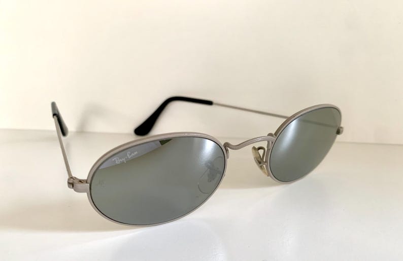 Puede incluir: Gafas de sol Ray-Ban plateadas con lentes ovaladas y acabado espejado. El logotipo de Ray-Ban es visible en la lente izquierda.