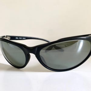 May include: Black sunglasses with a wraparound frame design and dark grey lenses. The inner temple arm features the white text B&L RAY BAN. These sunglasses have a curved, aerodynamic shape that provides full coverage around the eyes.