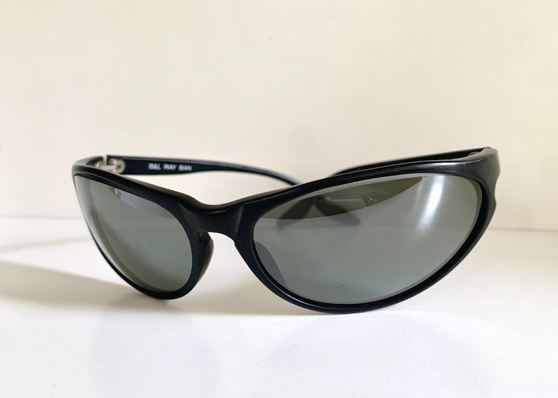 May include: Black sunglasses with a wraparound frame design and dark grey lenses. The inner temple arm features the white text B&L RAY BAN. These sunglasses have a curved, aerodynamic shape that provides full coverage around the eyes.