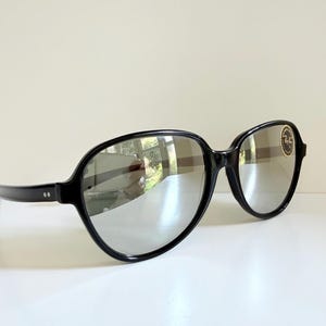 May include: Black-framed sunglasses with reflective lenses. The sunglasses have a rounded shape and a small, circular logo on the right lens. The frames are thick and glossy, and the lenses reflect the surrounding environment.