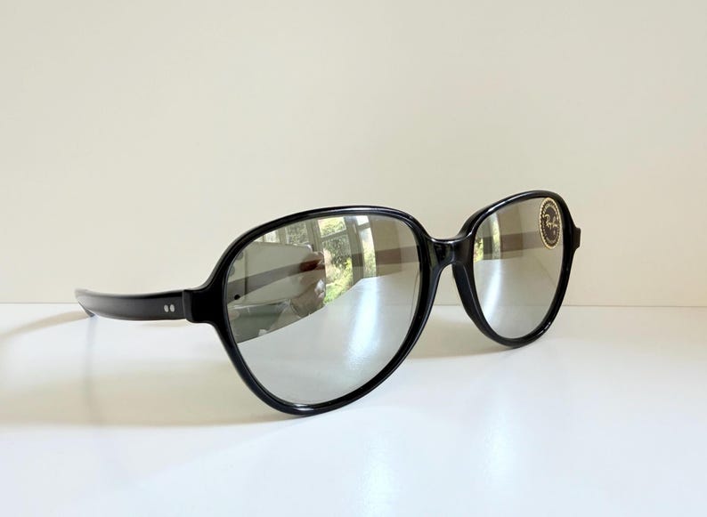 May include: Black-framed sunglasses with reflective lenses. The sunglasses have a rounded shape and a small, circular logo on the right lens. The frames are thick and glossy, and the lenses reflect the surrounding environment.