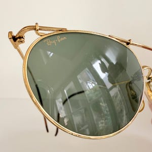 K&ouml;nnte beinhalten: Nahaufnahme einer Ray-Ban-Sonnenbrille mit goldenem Rahmen und gr&uuml;nen Gl&auml;sern. Das Ray-Ban-Logo ist auf dem Glas zu sehen. Die Sonnenbrille hat ein klassisches Design mit runder Form.
