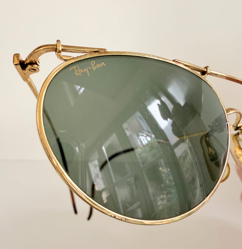 K&ouml;nnte beinhalten: Nahaufnahme einer Ray-Ban-Sonnenbrille mit goldenem Rahmen und gr&uuml;nen Gl&auml;sern. Das Ray-Ban-Logo ist auf dem Glas zu sehen. Die Sonnenbrille hat ein klassisches Design mit runder Form.