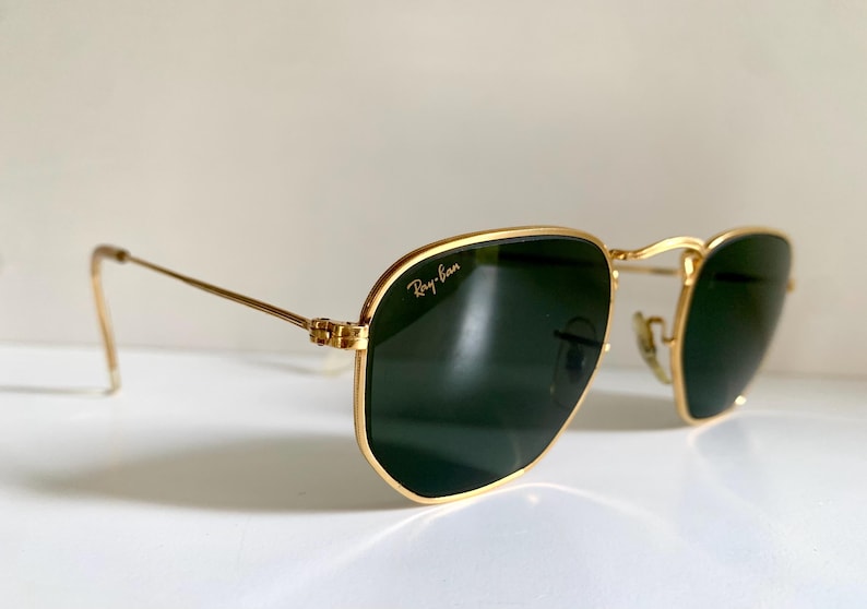 Może przedstawiać: Złote okulary przeciwsłoneczne z ciemnozielonymi soczewkami. Logo Ray-Ban jest widoczne na g&oacute;rnej lewej soczewce. Okulary mają klasyczny, kanciasty kształt i cienkie, złote zauszniki.