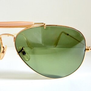 May include: Gold-framed aviator sunglasses with green lenses. The sunglasses have a double bridge and a light brown bar across the top. The lenses are teardrop-shaped and the frames are a shiny gold color.