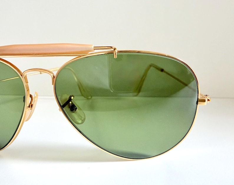 May include: Gold-framed aviator sunglasses with green lenses. The sunglasses have a double bridge and a light brown bar across the top. The lenses are teardrop-shaped and the frames are a shiny gold color.