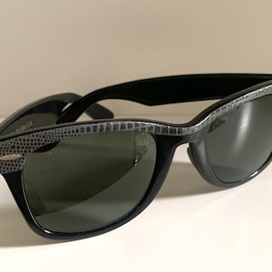 May include: Black sunglasses with a textured gray pattern on the top frame and temples. The lenses are a dark green color. The sunglasses are set against a white background.