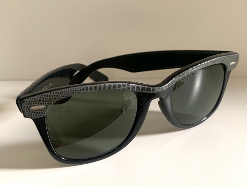 May include: Black sunglasses with a textured gray pattern on the top frame and temples. The lenses are a dark green color. The sunglasses are set against a white background.