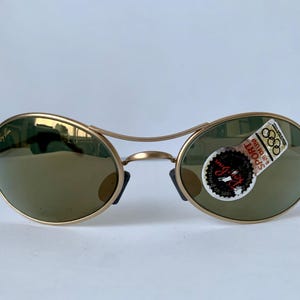 May include: Gold-tone Ray-Ban sunglasses with oval lenses and a double bridge. The lenses are a muted green color. A sticker on the right lens reads "SPORT" and "Ray-Ban".
