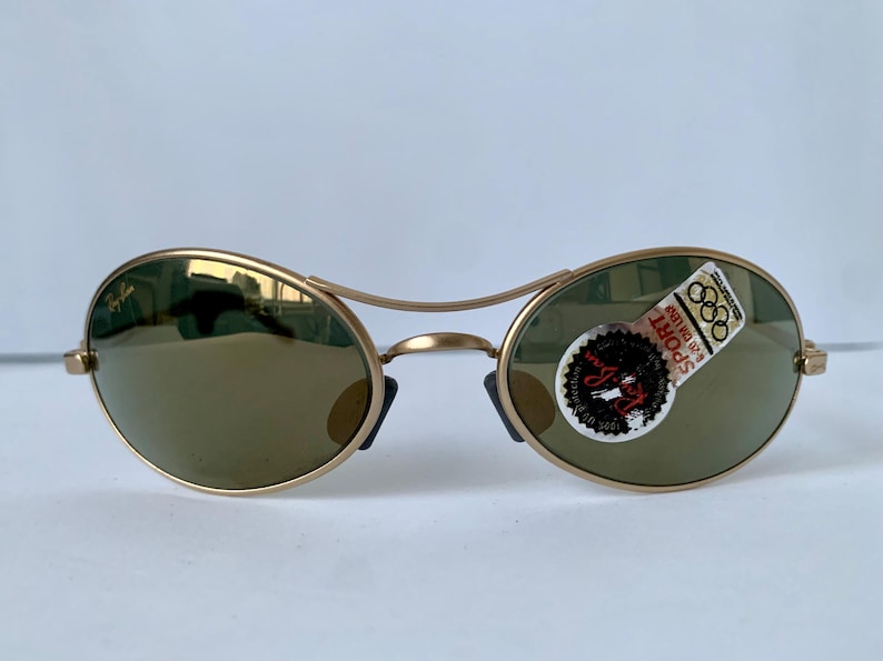 May include: Gold-tone Ray-Ban sunglasses with oval lenses and a double bridge. The lenses are a muted green color. A sticker on the right lens reads "SPORT" and "Ray-Ban".
