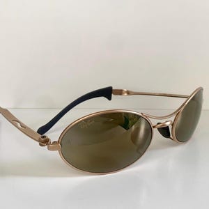 May include: Gold-tone oval sunglasses with brown lenses. The sunglasses have a sleek, modern design with black temple tips and the Ray-Ban logo on the lens. The frames are a light gold color.