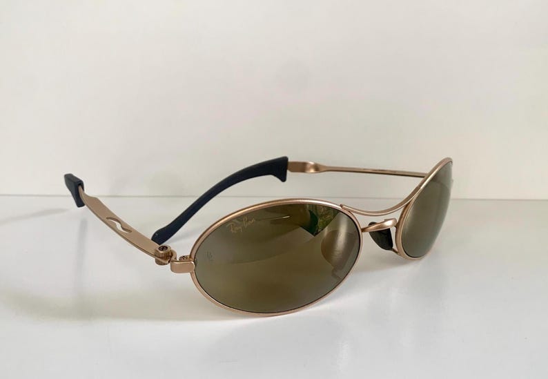 May include: Gold-tone oval sunglasses with brown lenses. The sunglasses have a sleek, modern design with black temple tips and the Ray-Ban logo on the lens. The frames are a light gold color.