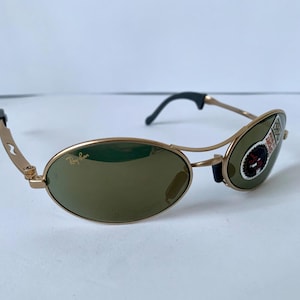 May include: Gold-framed sunglasses with oval lenses and a dark tint. The sunglasses have a small logo on the lens and a sticker on the right lens. The arms are gold with black ear pieces.