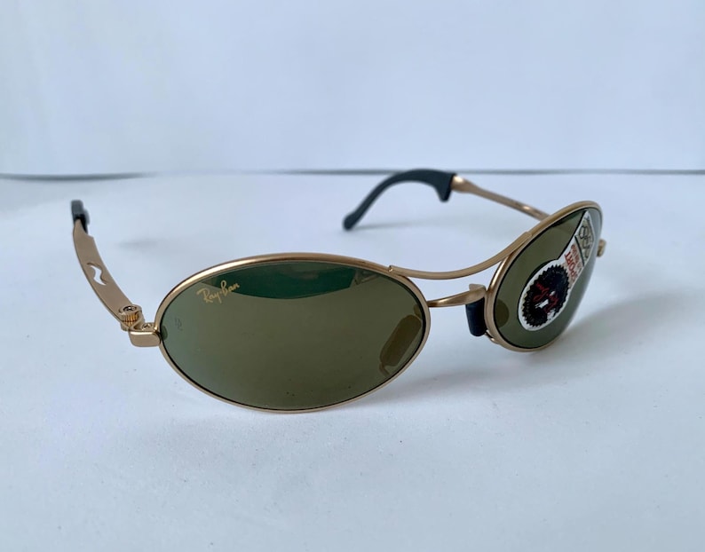 May include: Gold-framed sunglasses with oval lenses and a dark tint. The sunglasses have a small logo on the lens and a sticker on the right lens. The arms are gold with black ear pieces.