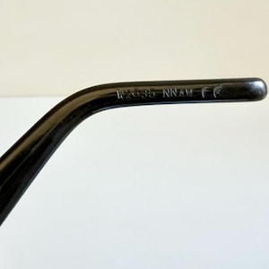 May include: A close-up of a black spectacle arm with the text "17635 NNAM CE" printed on it. The arm is curved and has a glossy finish. The background is a plain white surface.