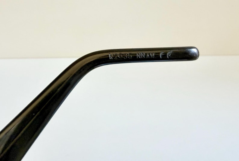 May include: A close-up of a black spectacle arm with the text "17635 NNAM CE" printed on it. The arm is curved and has a glossy finish. The background is a plain white surface.