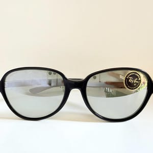 May include: Black sunglasses with silver mirrored lenses. The sunglasses have a thick, rounded frame. A gold and black sticker with the Ray-Ban logo is on the right lens. The sunglasses are set against a white background.