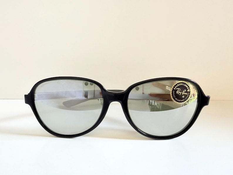 May include: Black sunglasses with silver mirrored lenses. The sunglasses have a thick, rounded frame. A gold and black sticker with the Ray-Ban logo is on the right lens. The sunglasses are set against a white background.