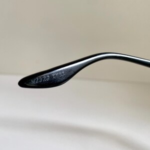 May include: A black eyeglass temple arm featuring a curved design and a smooth, glossy finish. The interior side of the temple arm is clearly embossed with the alphanumeric text W2323 XVAS, which is visible in a crisp, legible font style.