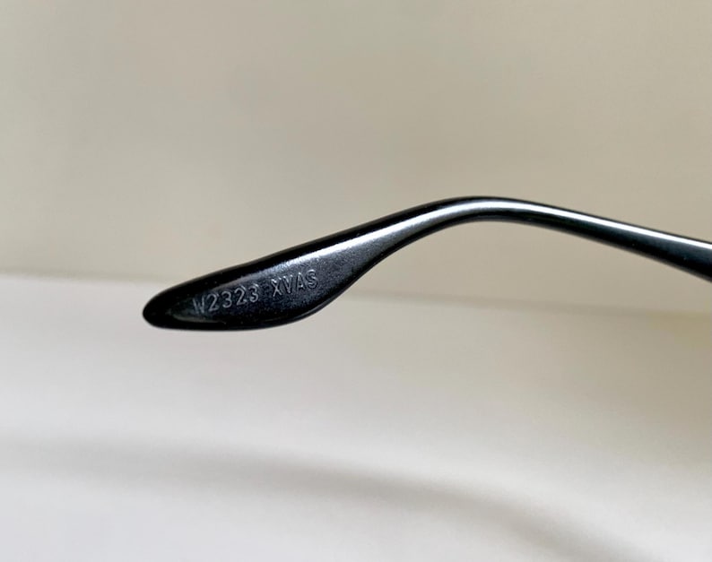 May include: A black eyeglass temple arm featuring a curved design and a smooth, glossy finish. The interior side of the temple arm is clearly embossed with the alphanumeric text W2323 XVAS, which is visible in a crisp, legible font style.