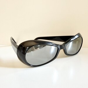 May include: Black sunglasses with a thick, rounded frame and reflective lenses. The sunglasses have a sleek, modern design. The lenses reflect the surrounding environment, adding a touch of mystery. Suitable for various styles.