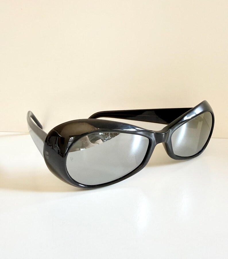 May include: Black sunglasses with a thick, rounded frame and reflective lenses. The sunglasses have a sleek, modern design. The lenses reflect the surrounding environment, adding a touch of mystery. Suitable for various styles.